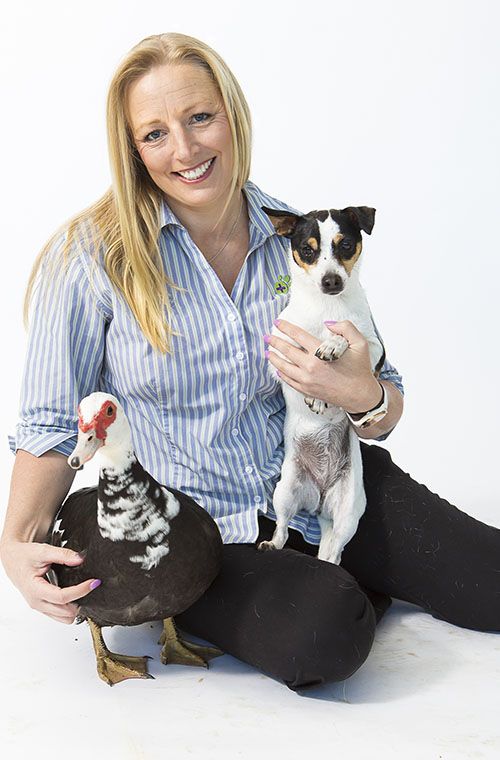 Woman sitting with a small dog and a duck, smiling at the camera.