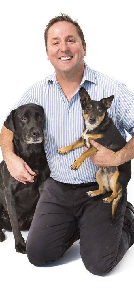 Man kneeling with a black Labrador and a small mixed-breed dog.